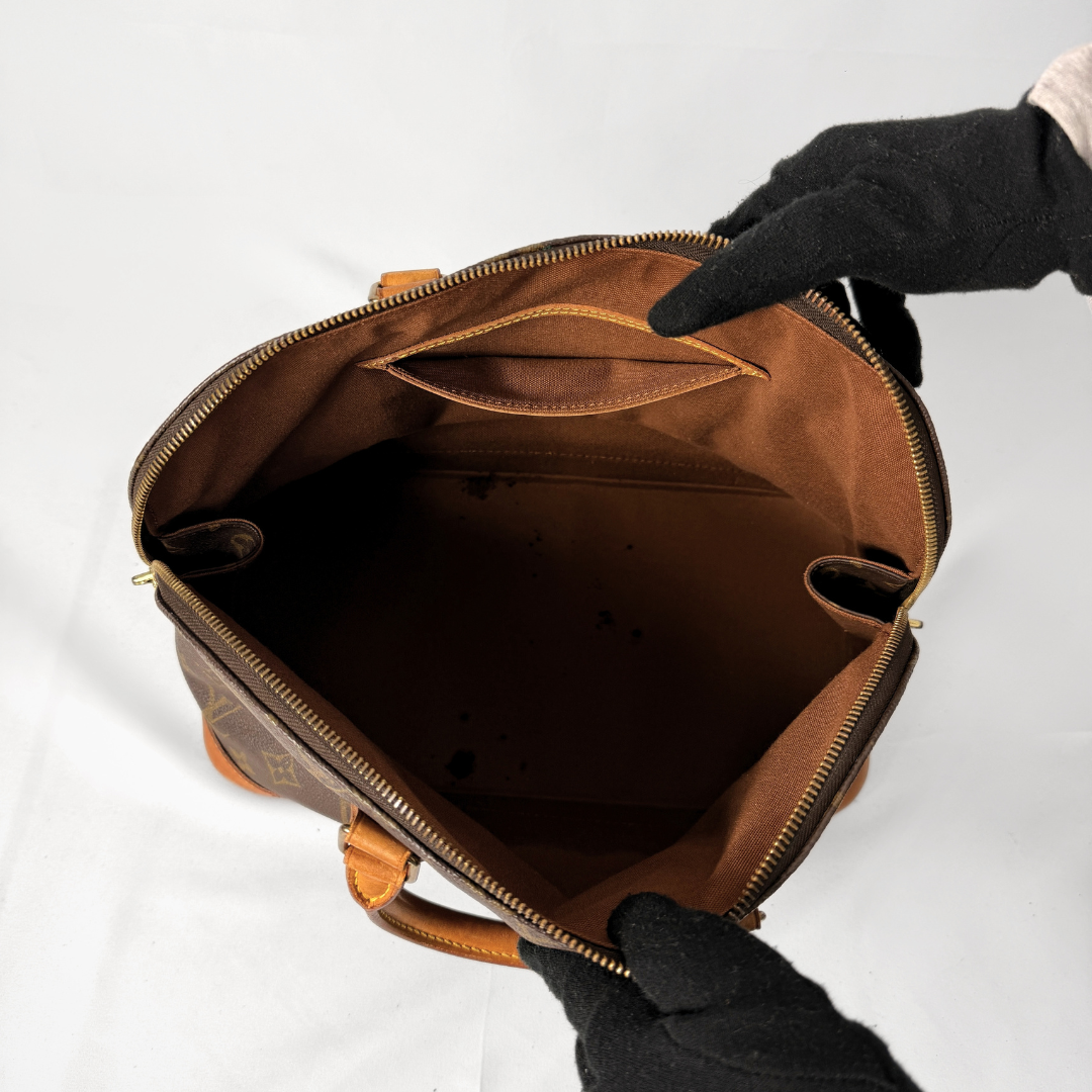 A brown monogram handbag shown open, with black gloved hands holding it against a white background to display the interior.