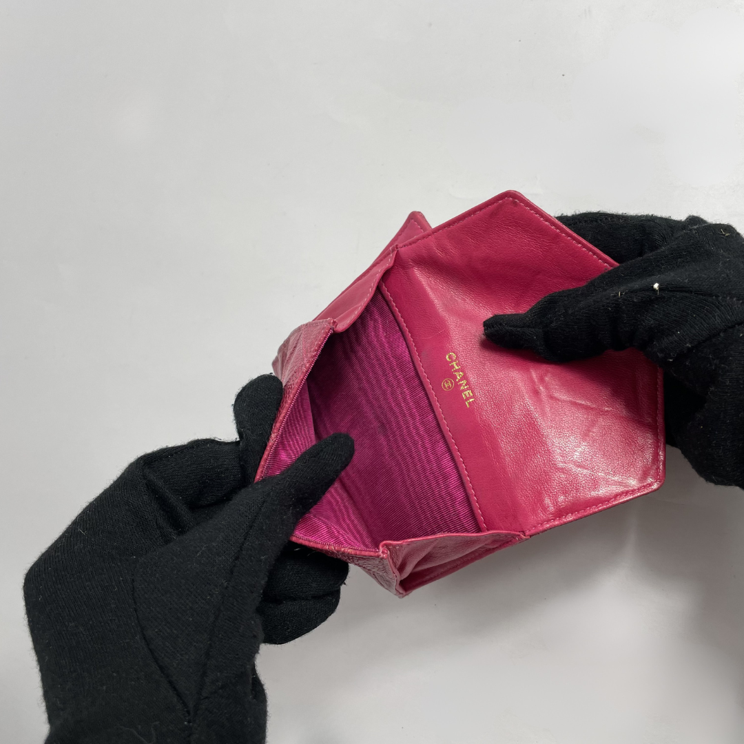A pink Chanel wallet is shown open, held by hands wearing black gloves. The brand name 'CHANEL' is printed in gold on the interior flap.
