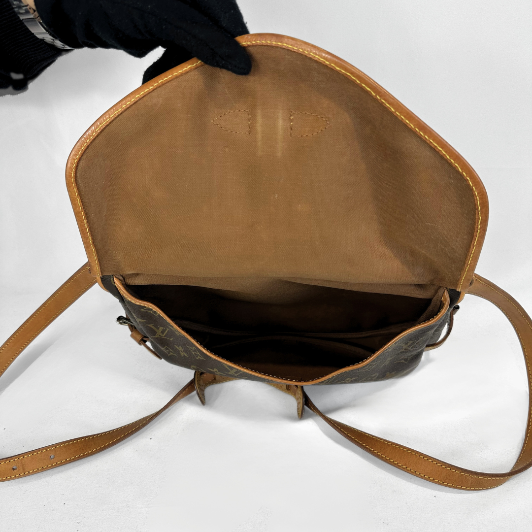 A Louis Vuitton monogram shoulder bag shown open, displaying its tan leather interior and lining. A hand wearing a black glove holds the top edge of the bag, which is set against a plain white background.