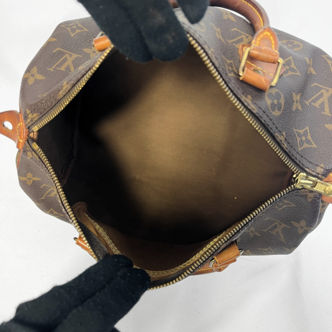 An open Louis Vuitton monogram handbag shown from above, with black-gloved hands holding the opening wide to display the interior. The bag features the classic brown coated canvas with tan monogram motifs, tan leather trim, and gold-tone zippers.