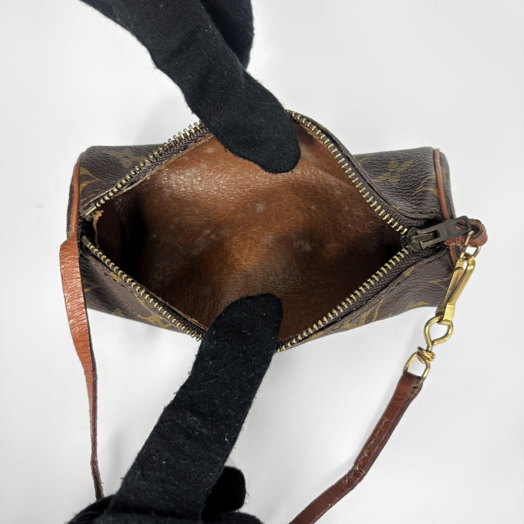 A close-up of a Louis Vuitton monogram canvas pouch with a brown leather interior, shown open with a black gloved hand pointing inside. The gold-tone clasp and brown leather strap are visible against a plain white background.