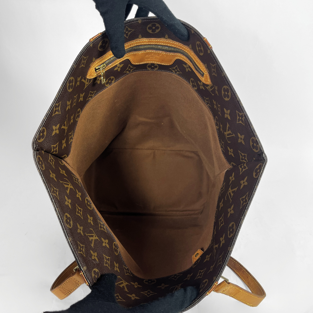 An open Louis Vuitton monogram canvas tote bag, showing the brown interior lining and tan leather trim, held open by black gloves against a white background.