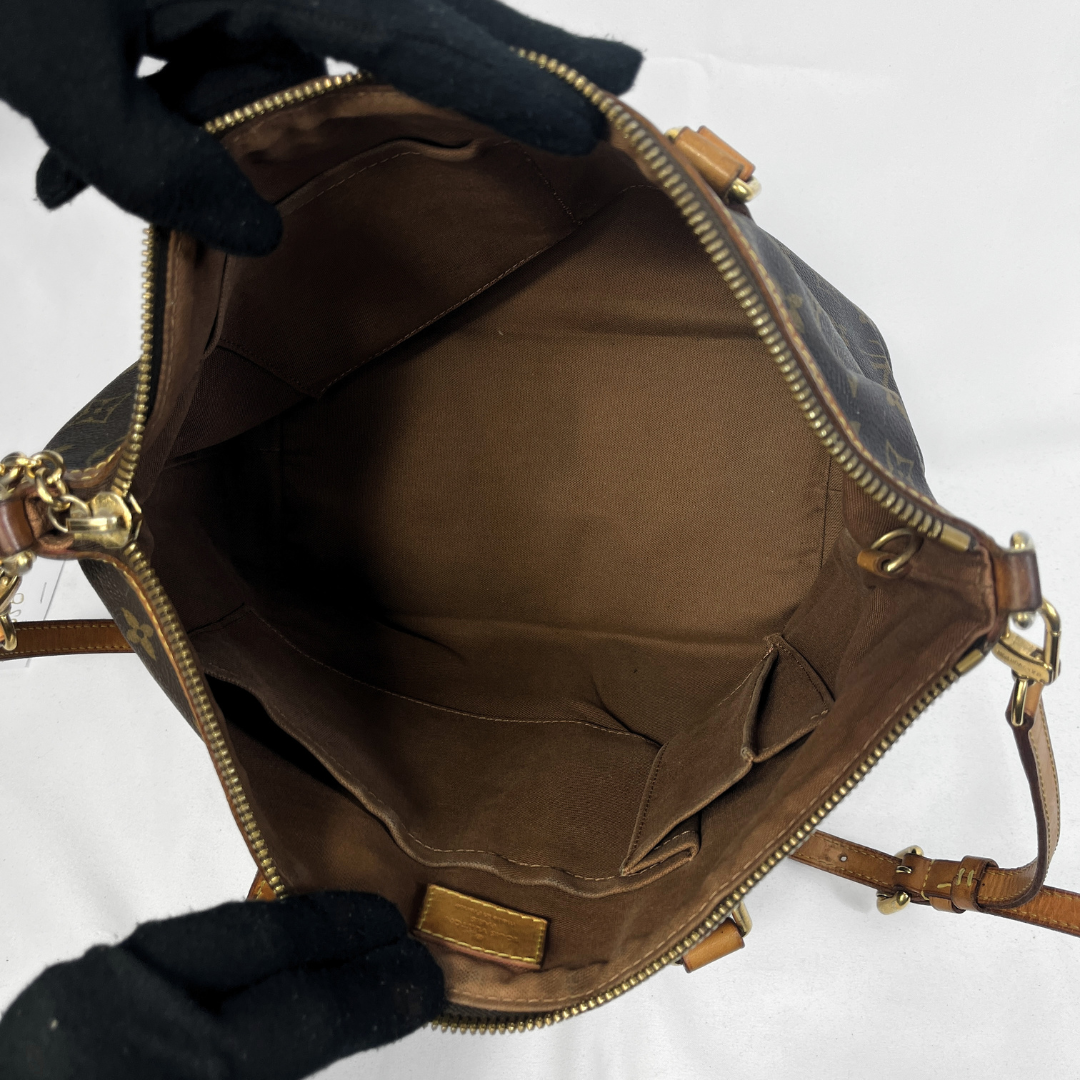 An open Louis Vuitton shoulder bag shown from the inside, featuring a brown monogram canvas exterior, a brown fabric lining, and gold-tone hardware. Black gloves are holding the bag open against a white background.