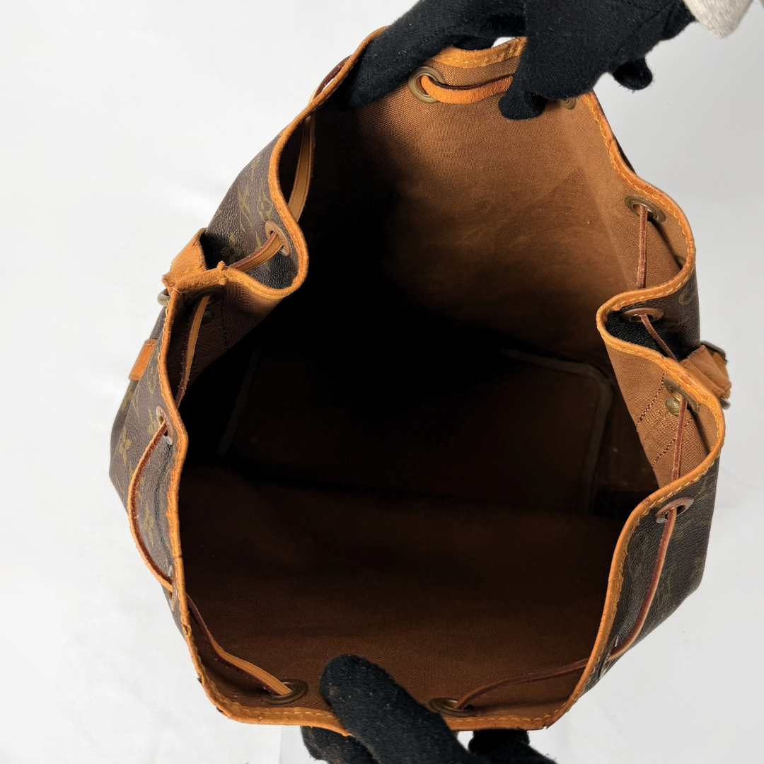 An open brown monogram canvas bucket bag with tan leather trim, shown from above against a white background, with gloved hands holding the opening.