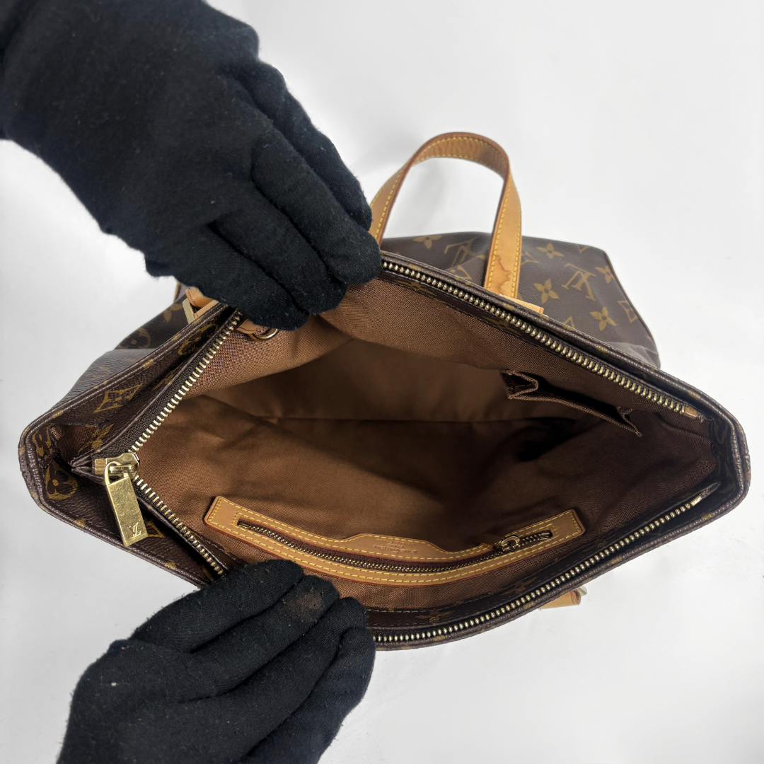 A Louis Vuitton monogram handbag shown open from above, with black gloves holding the top and sides to display the interior. The bag has a brown coated canvas body with the classic tan monogram print, tan leather handles, and a tan interior lining with gold-tone zippers.