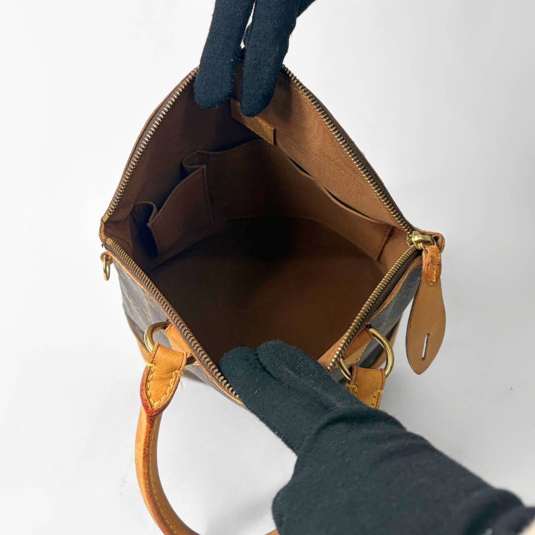 Brown leather handbag with gold accents being held open by a gloved hand on a white background