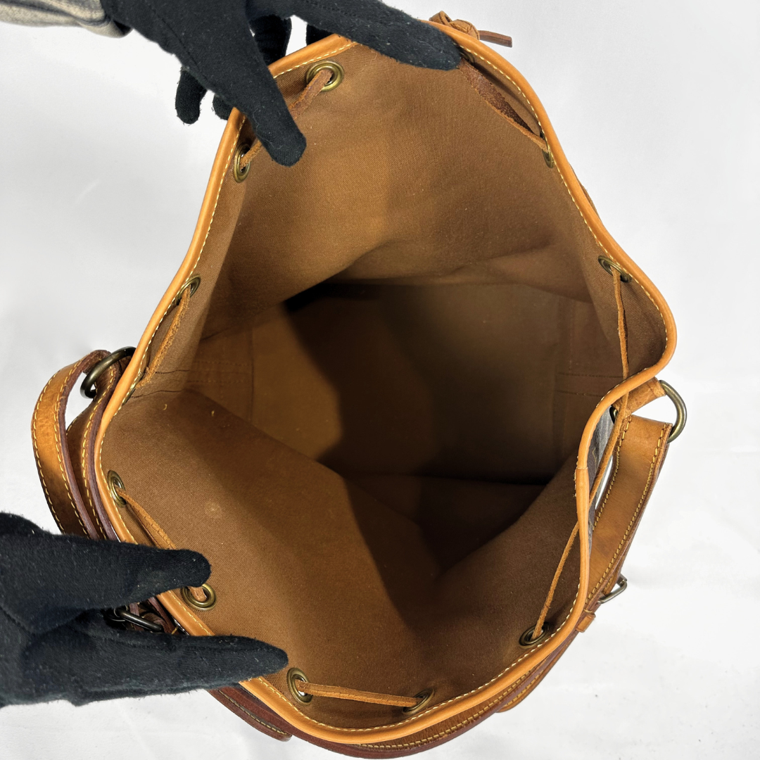 A top-down view of a tan leather bucket bag being held open by two hands wearing black gloves, showing the interior lining and brass grommets against a white background.