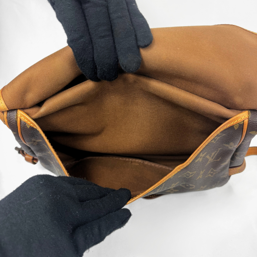 A Louis Vuitton monogram canvas shoulder bag in brown and tan, shown open with black gloves holding it to display the interior lining.
