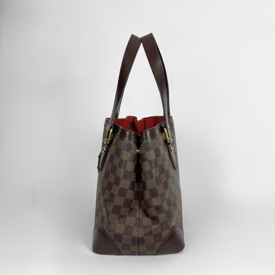 A Louis Vuitton Damier Ebene canvas tote bag with dark brown leather handles and trim, featuring a red interior lining, set against a plain light gray background.