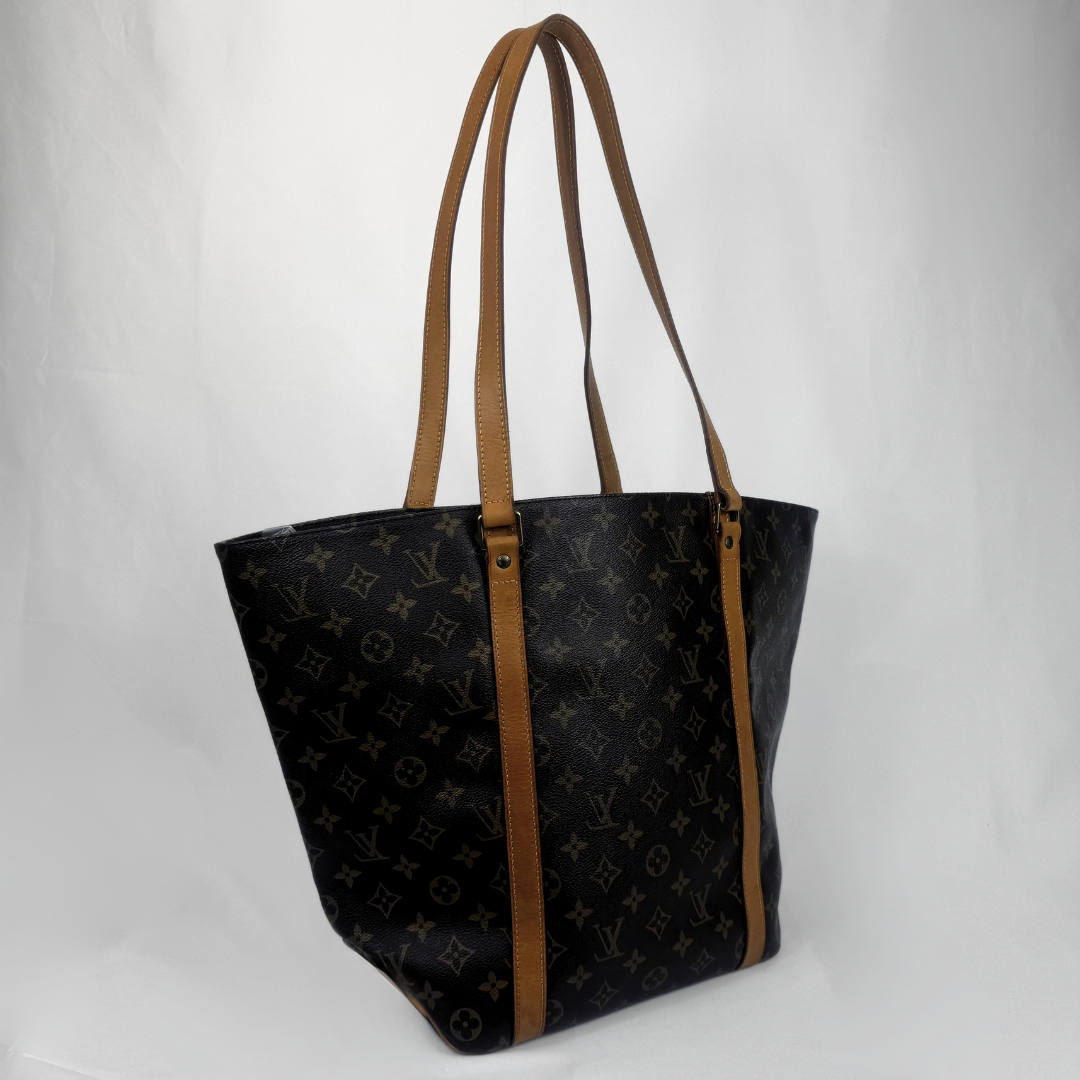 A Louis Vuitton tote bag in black monogram canvas with brown leather shoulder straps and trim, photographed against a plain light gray background.