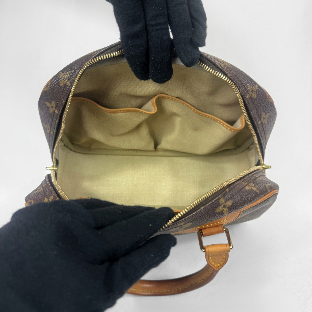 A Louis Vuitton monogram handbag shown open, with black gloves holding the zipper area to display the interior lining and compartments. The bag features the classic brown and beige monogram pattern, tan leather trim, and gold-tone hardware on a plain white background.