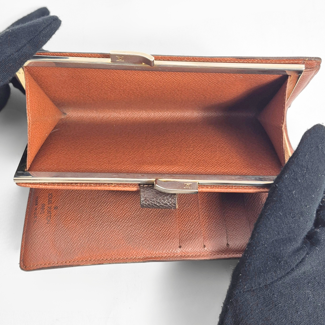 Bolso de mano de piel marrón con detalles dorados, sostenido por una mano enguantada sobre un fondo gris claro.