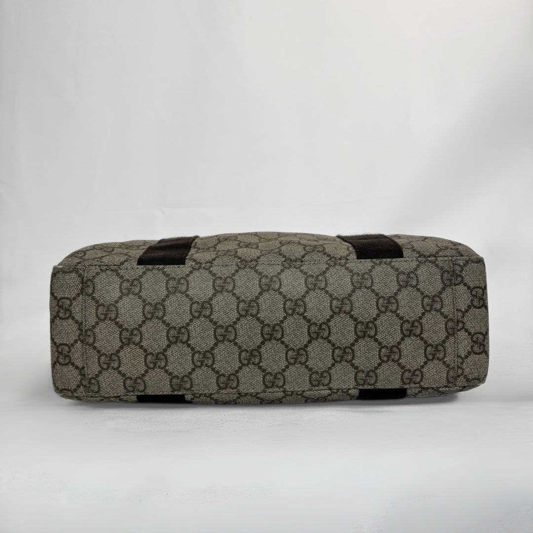 A cylindrical canvas bag featuring the classic brown and beige GG monogram pattern, with dark brown leather accents on the top handles and bottom edges, photographed against a neutral light gray background.