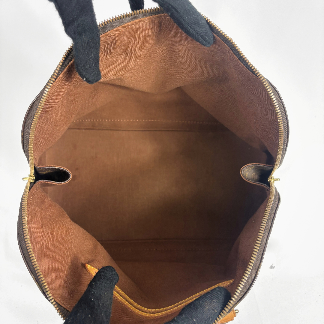 Top-down view of an open brown leather handbag with a tan suede-like interior, gold-tone zipper, and black handles against a white background.