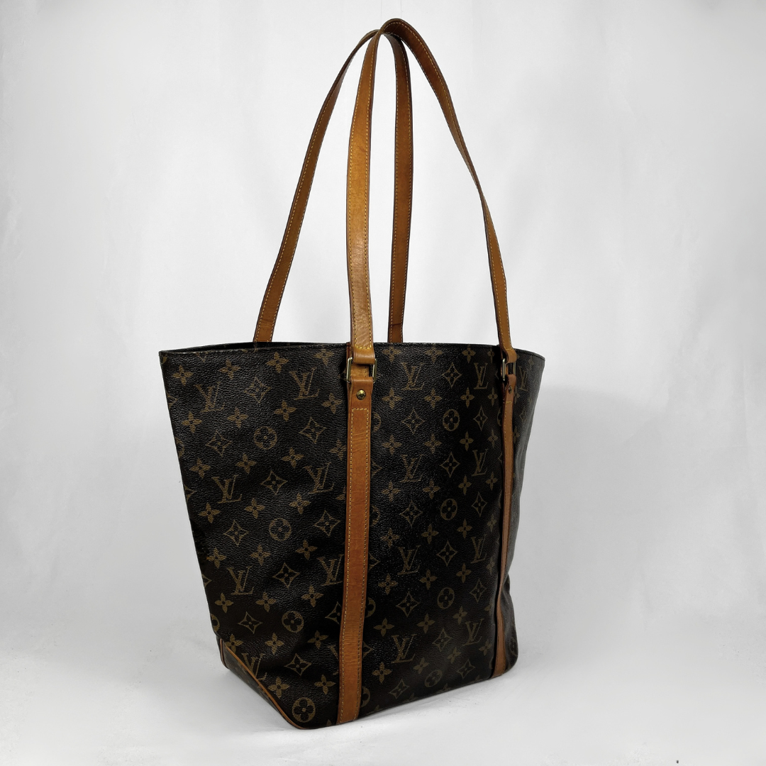 A Louis Vuitton tote bag in brown monogram canvas with tan leather shoulder straps and trim, photographed against a plain light gray background.