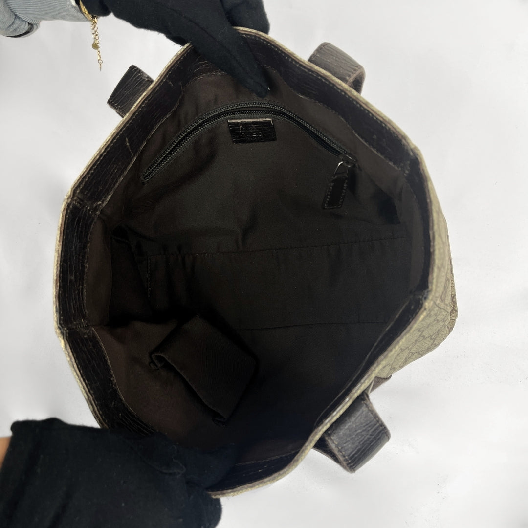 An open tote bag shown from above, revealing a dark brown interior with a zip pocket and fabric lining. A black gloved hand holds the top edge of the bag against a plain white background.