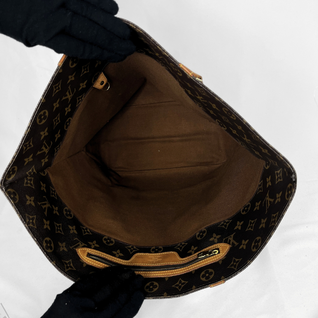Louis Vuitton monogram canvas handbag shown open from the outside, with black-gloved hands holding the top and zipper area against a plain white background.