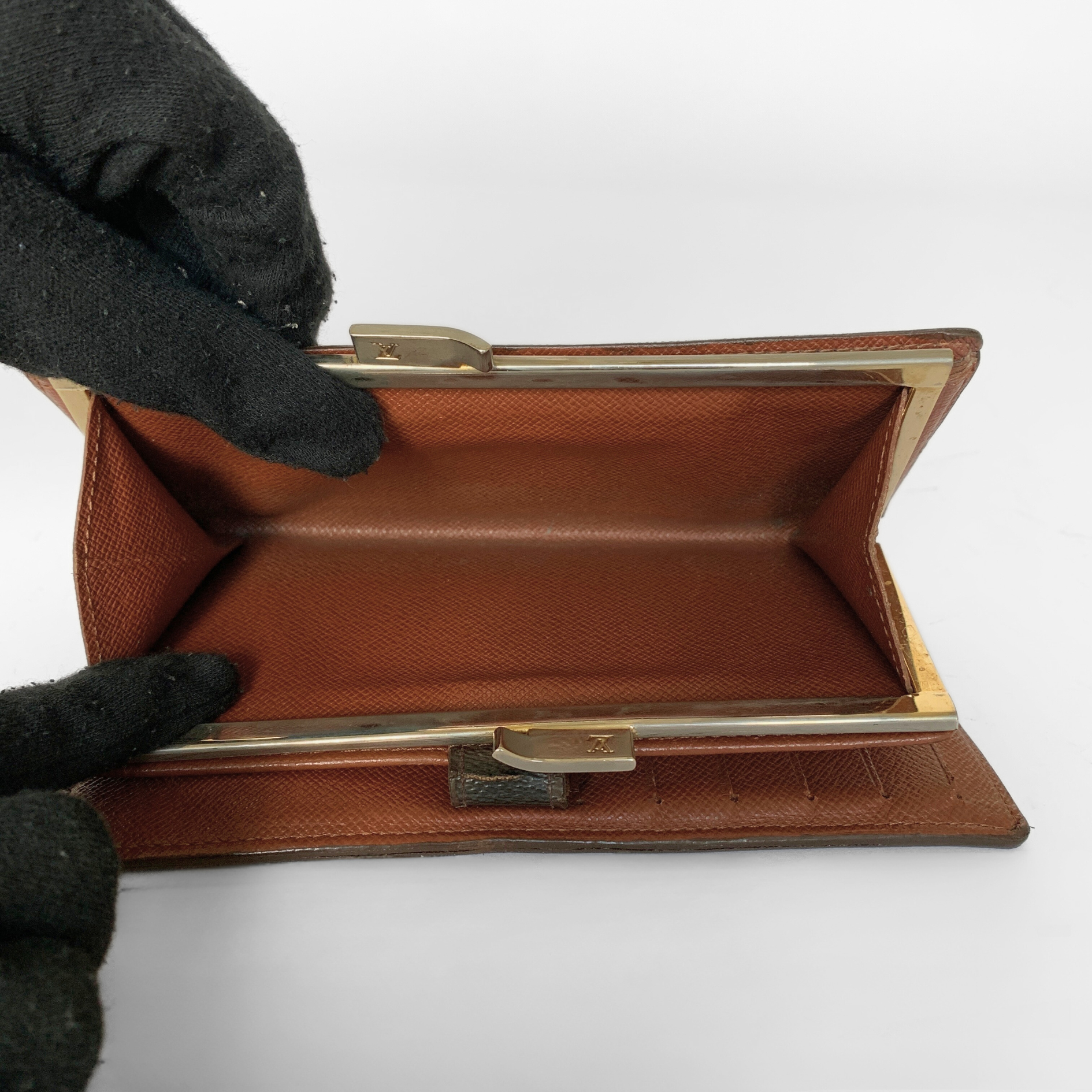 A brown textured leather clutch wallet with a gold-tone metal frame, shown open with a black-gloved hand holding it against a white background.