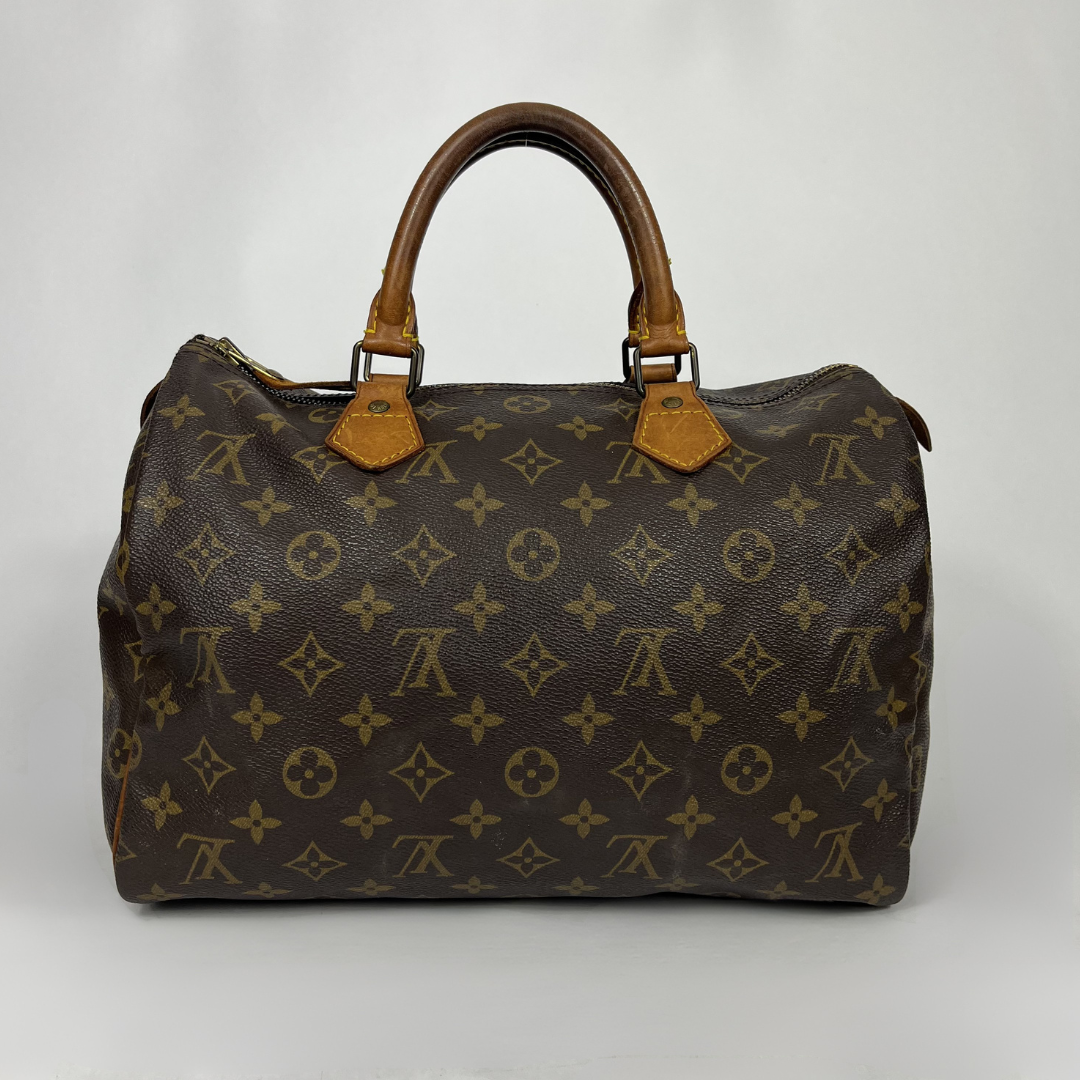 A Louis Vuitton monogram canvas speedy bag with brown leather handles and gold-tone hardware, photographed against a neutral light gray background.