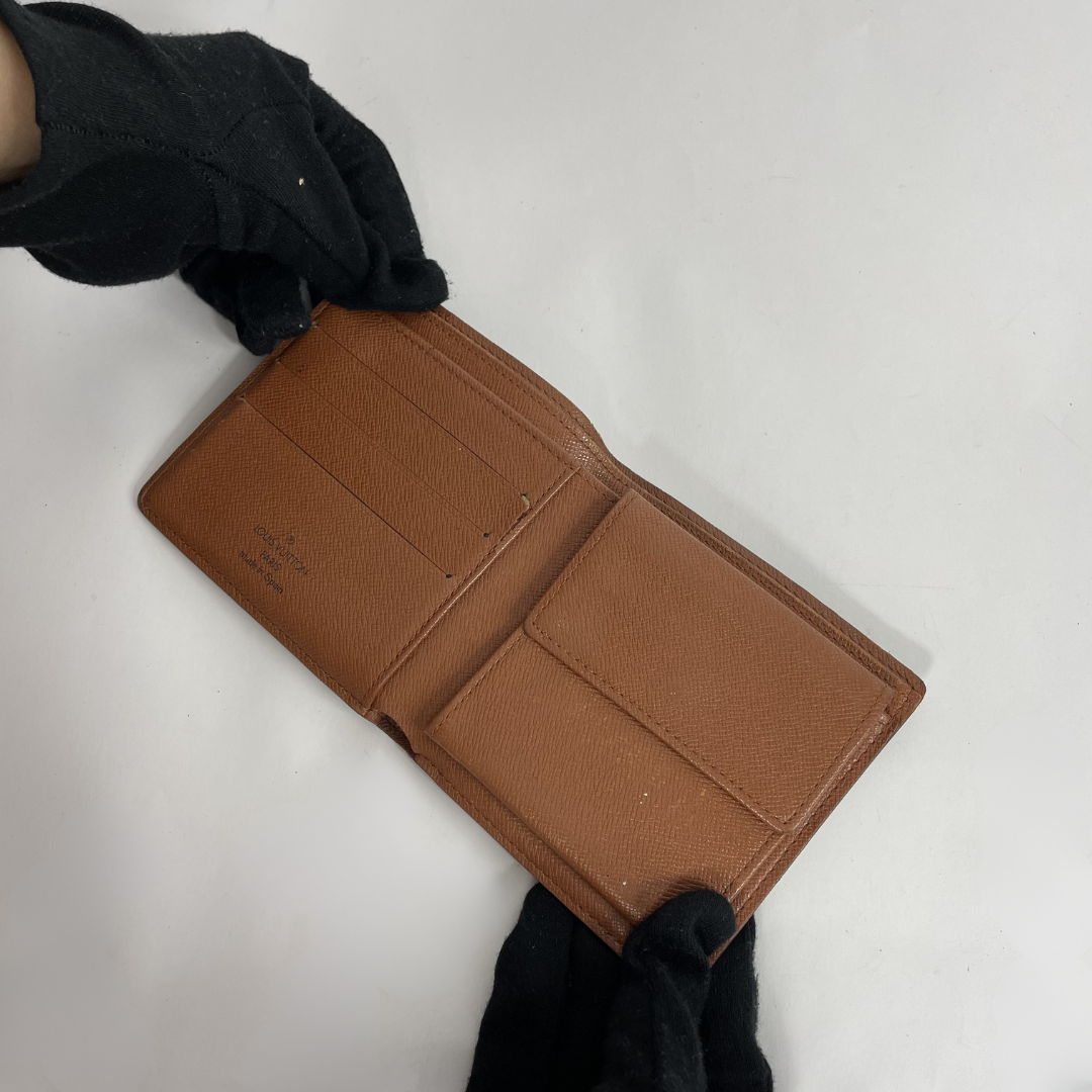 A brown textured leather bifold wallet shown open, held by hands wearing black gloves on a plain white background.