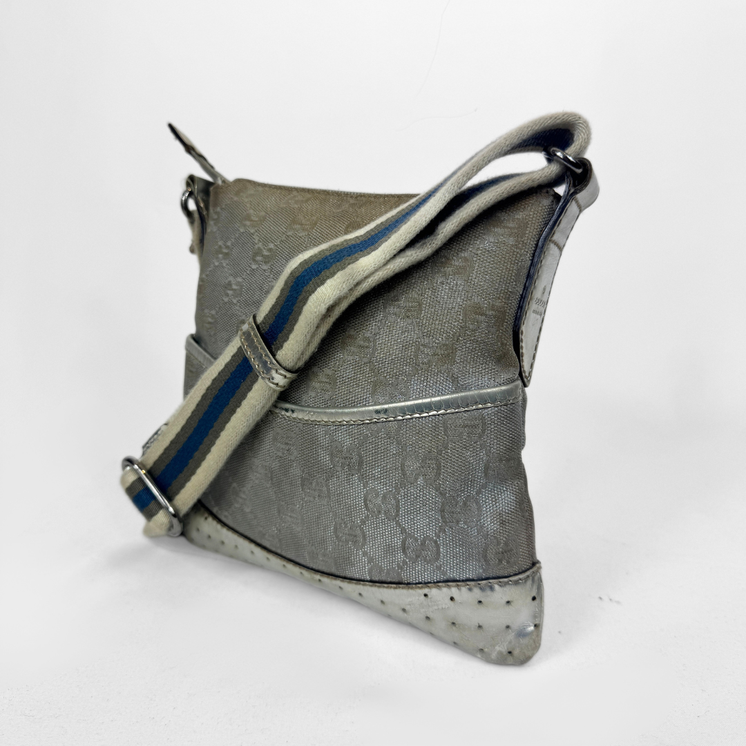 A Gucci GG canvas crossbody bag in metallic silver with a blue and beige striped shoulder strap, silver-tone hardware and a studded base, photographed against a plain white background.