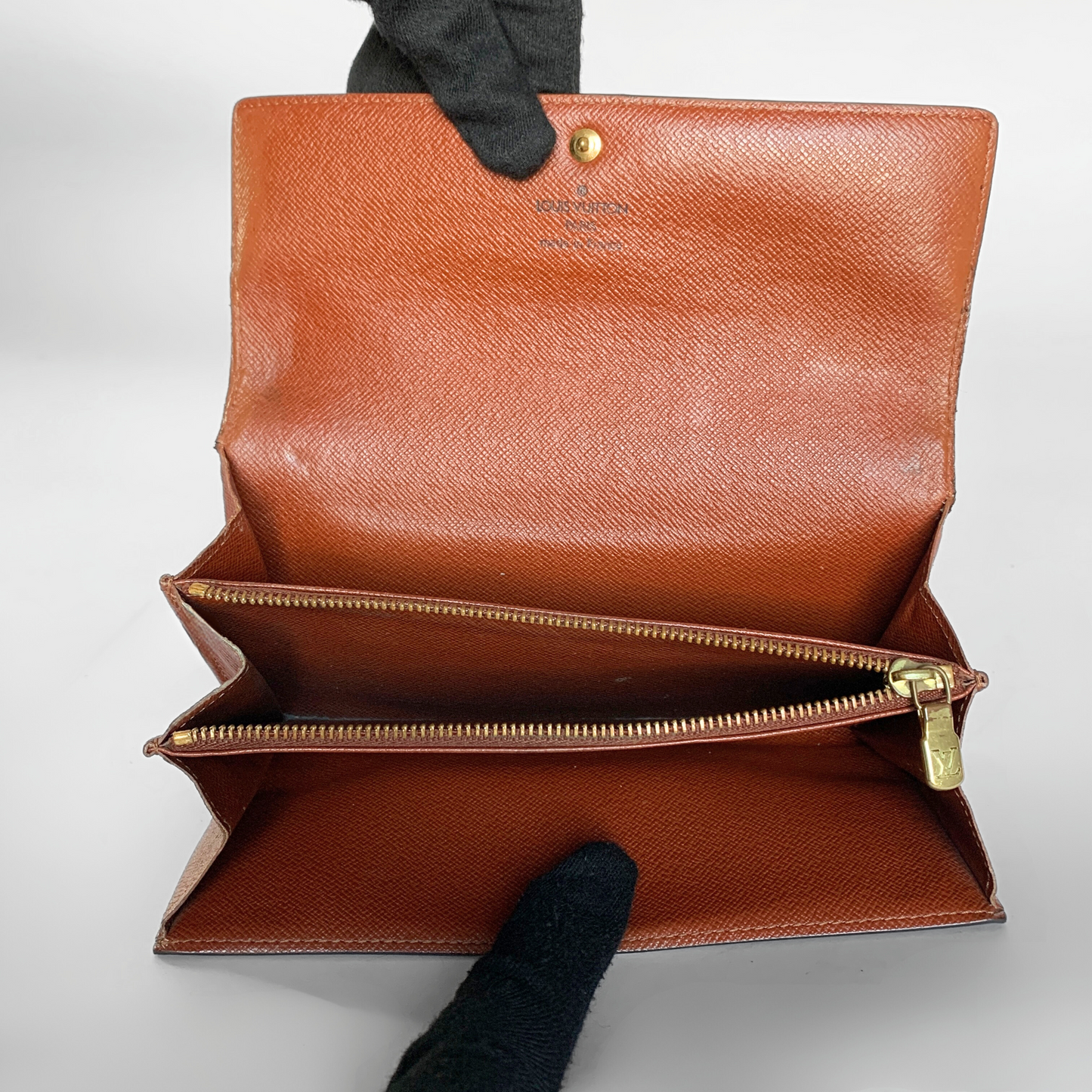 An open brown leather wallet or clutch shown from the inside, with gold-tone zippers and a small gold stud detail. Gloved hands are holding it open against a plain light background.