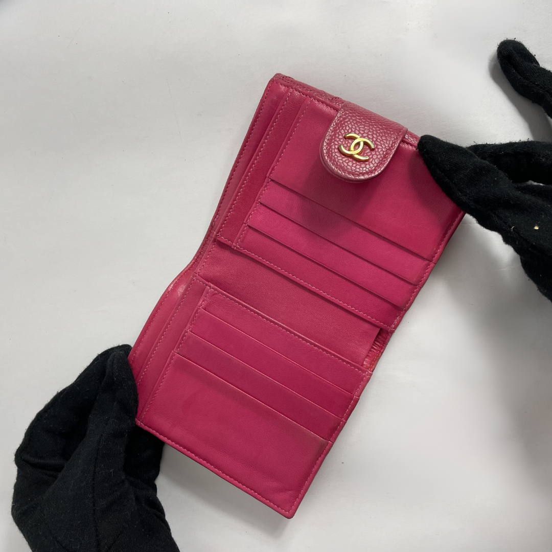 A bright pink Chanel wallet with a gold interlocking CC logo, held open by two hands wearing black gloves on a light gray background.