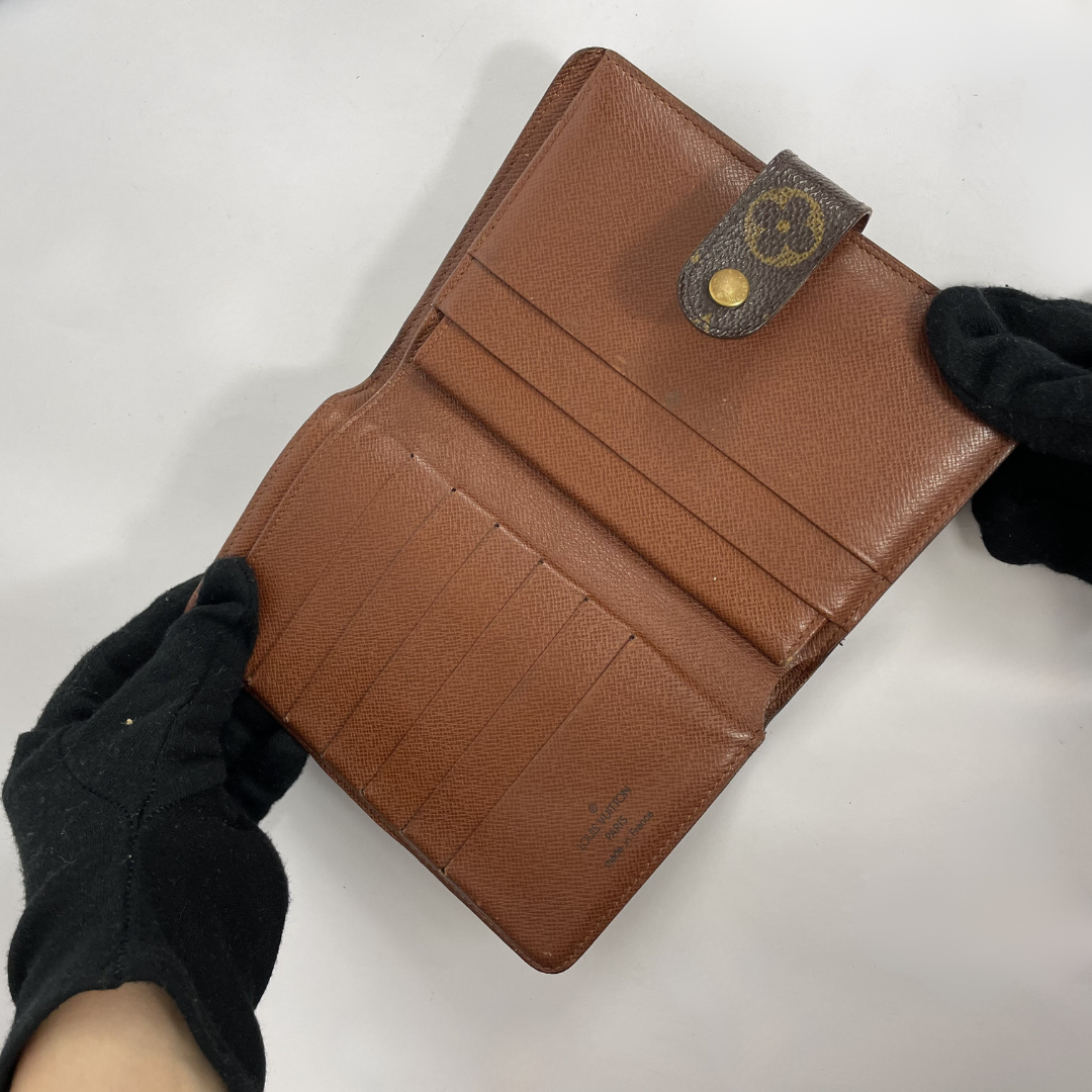 A brown pebbled leather wallet held open by hands wearing black gloves, showing its interior card slots and a snap-tab closure with a circular logo.