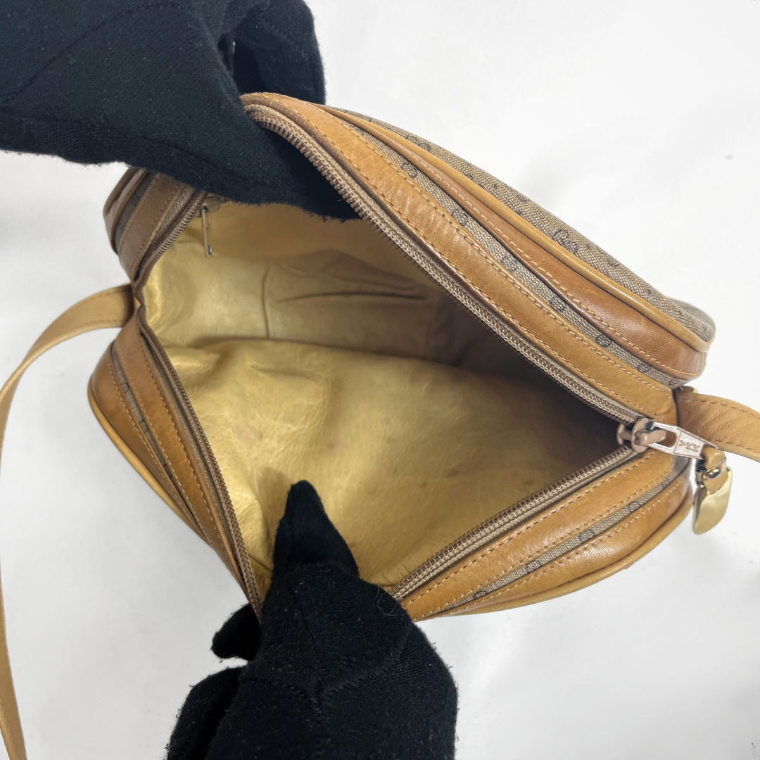 A tan leather shoulder bag with a gold interior lining, shown open with black-gloved hands holding it against a white background.