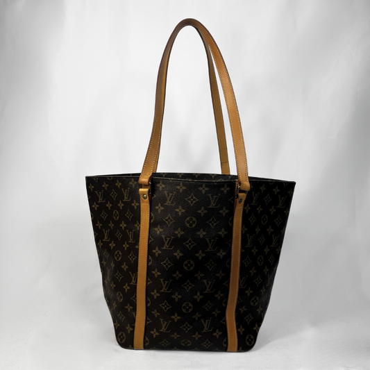 A Louis Vuitton tote bag in black monogram canvas with tan leather shoulder straps and trim, photographed against a light gray studio background.