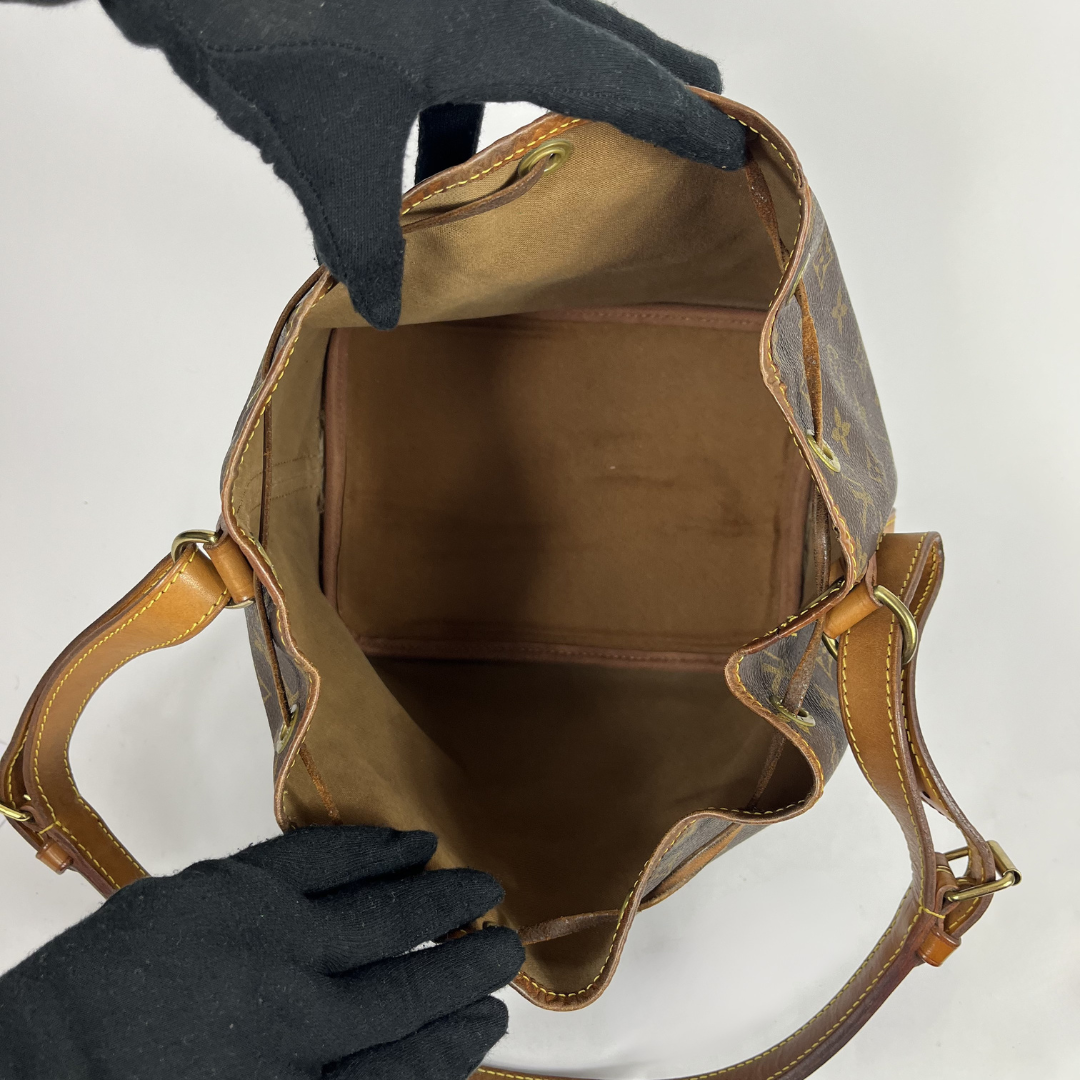 A Louis Vuitton monogram bucket bag shown open, with black gloved hands holding the top and sides to display the interior. The bag features classic brown monogram canvas, tan leather trim, and gold-tone hardware on a white background.
