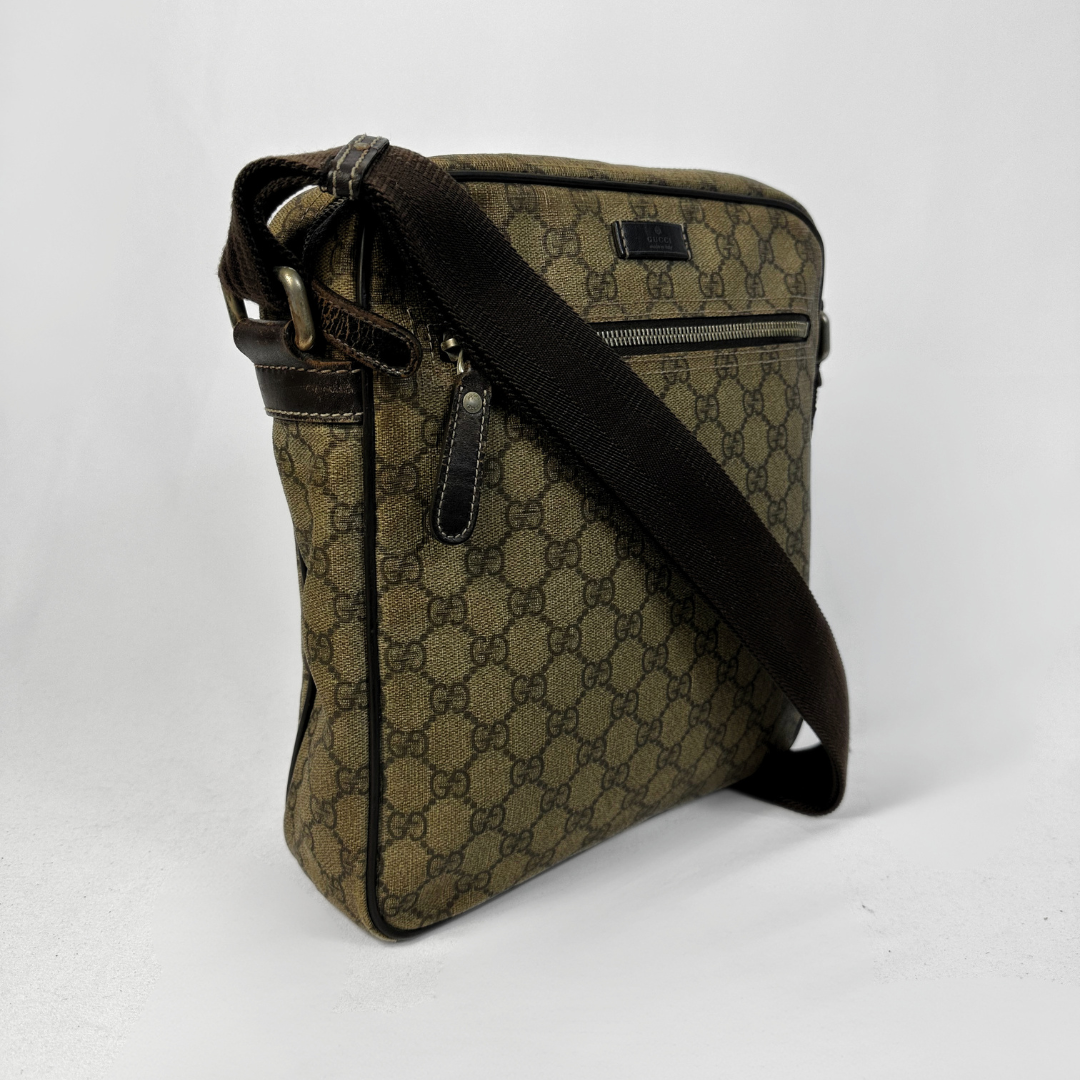 A vintage Gucci monogram canvas shoulder bag in brown and beige, featuring a black webbed shoulder strap and dark brown leather trim, photographed on a light gray background.