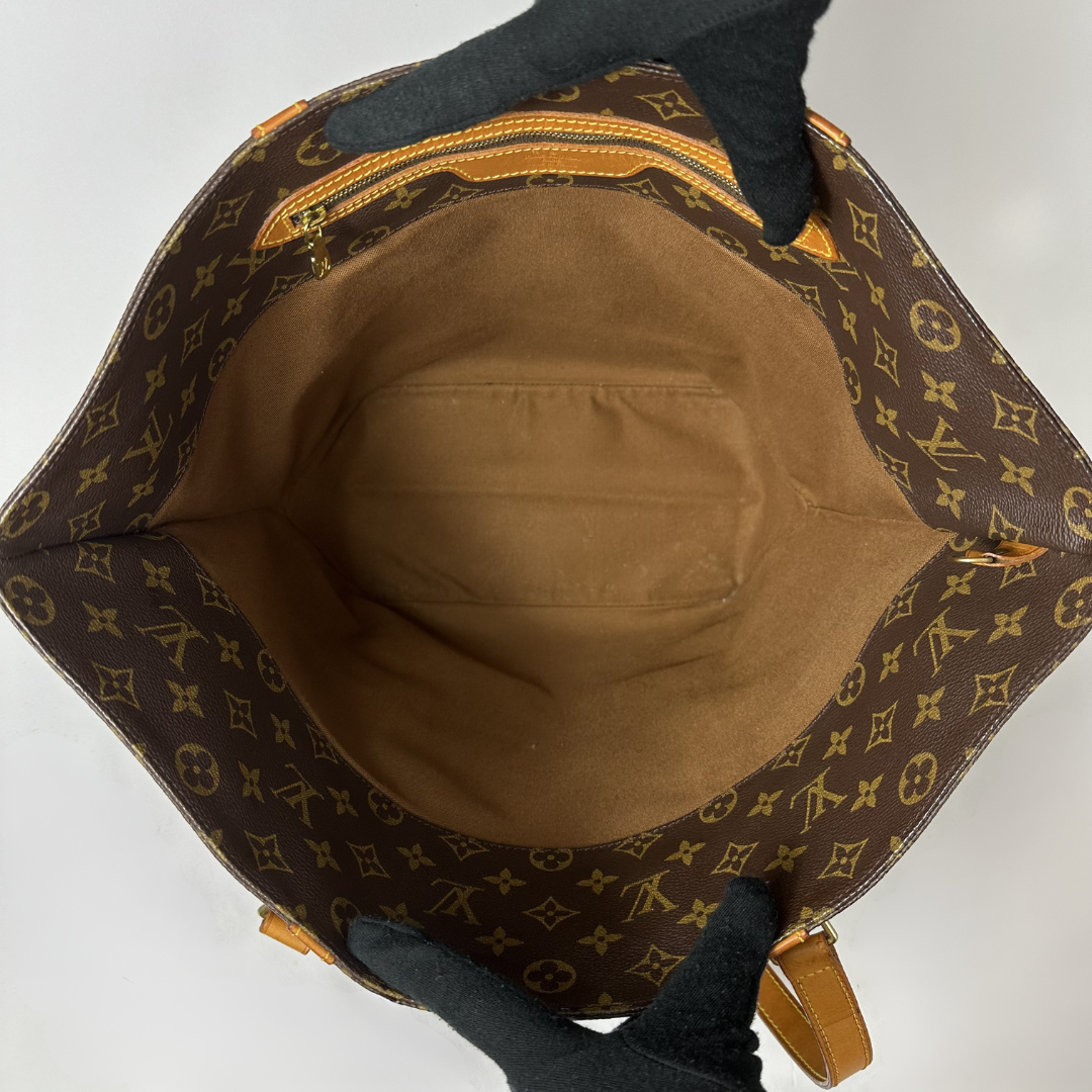 The interior of a Louis Vuitton monogram canvas tote bag, showing the brown interior lining and gold-tone zipper, held open by black gloves against a white background.