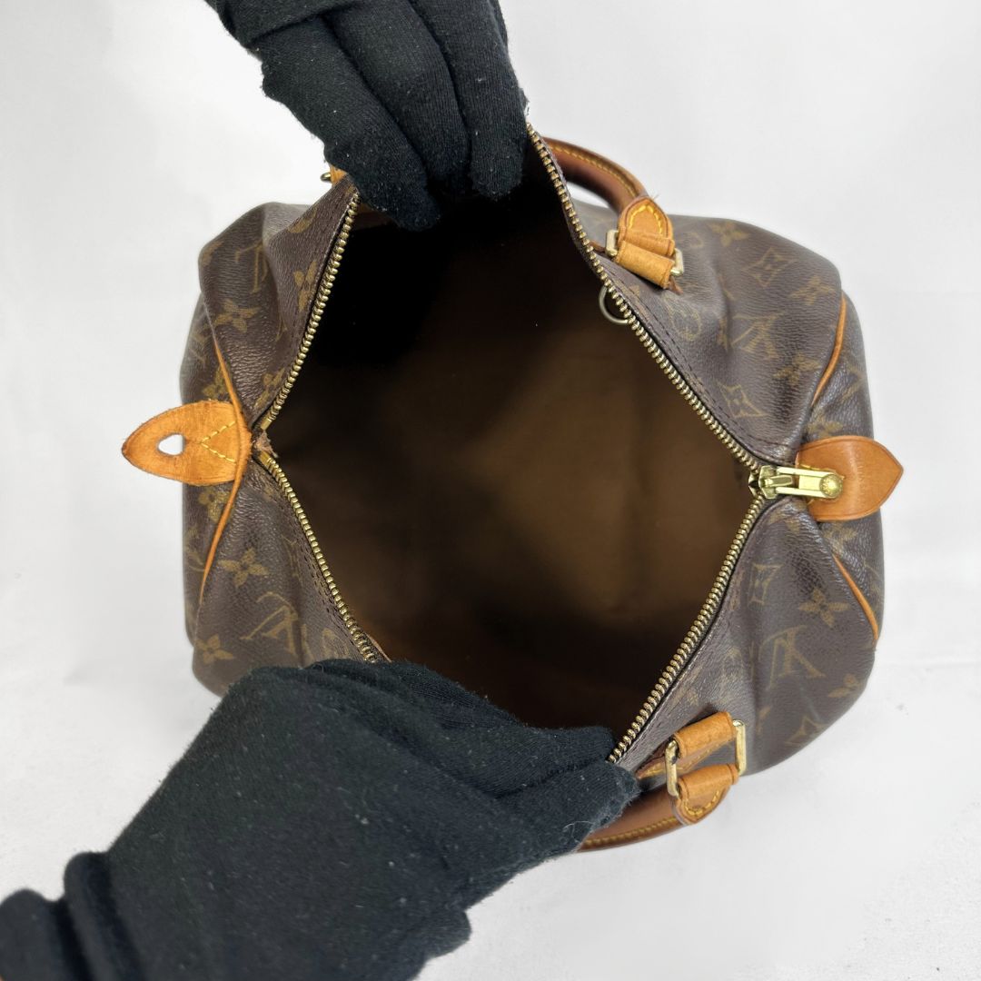 A Louis Vuitton monogram handbag shown open from above, with black-gloved hands holding the top zipper area. The bag has a brown coated-canvas body with tan leather trim and gold-tone hardware on a plain light background.
