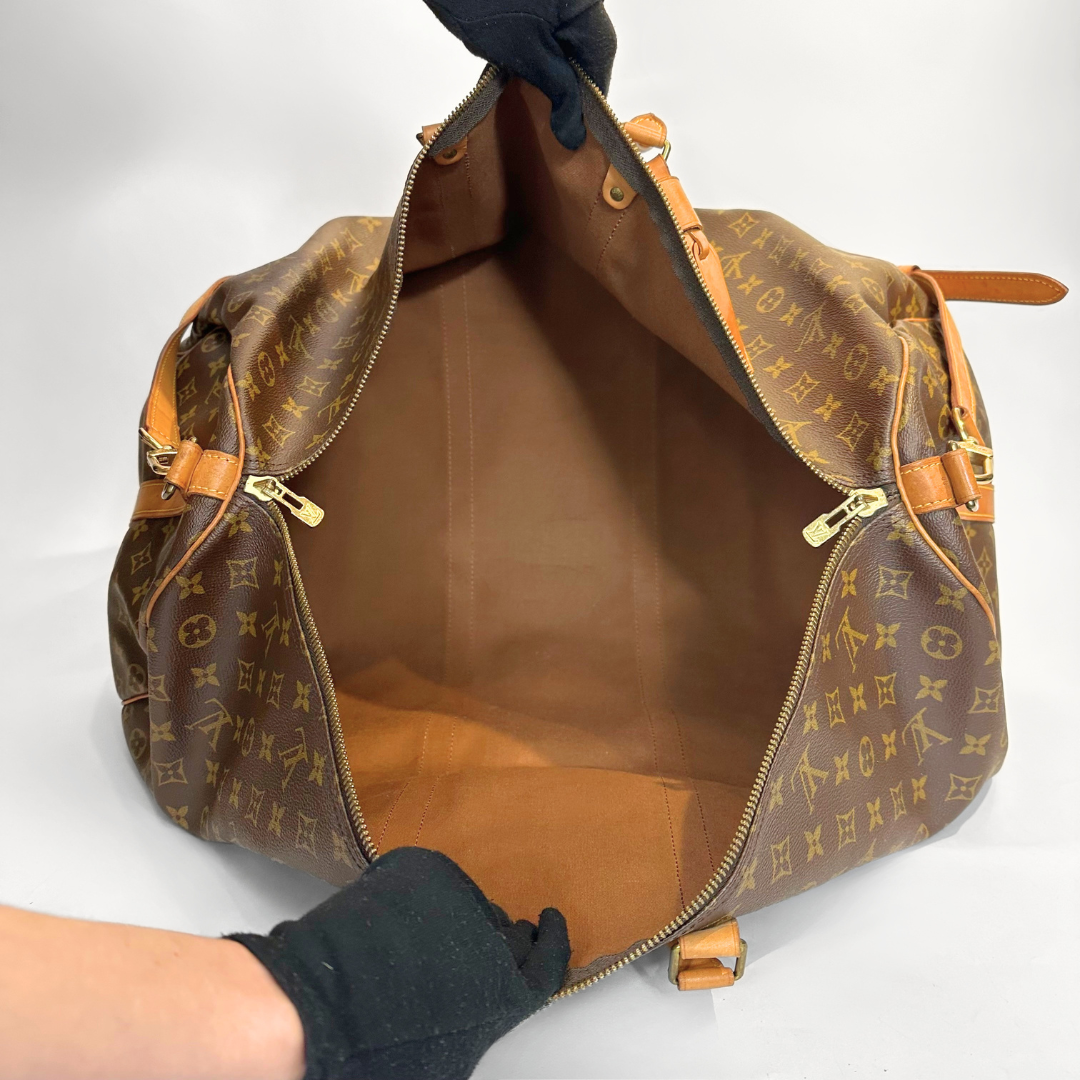 Brown monogrammed handbag with gold accents held by a gloved hand on a white background