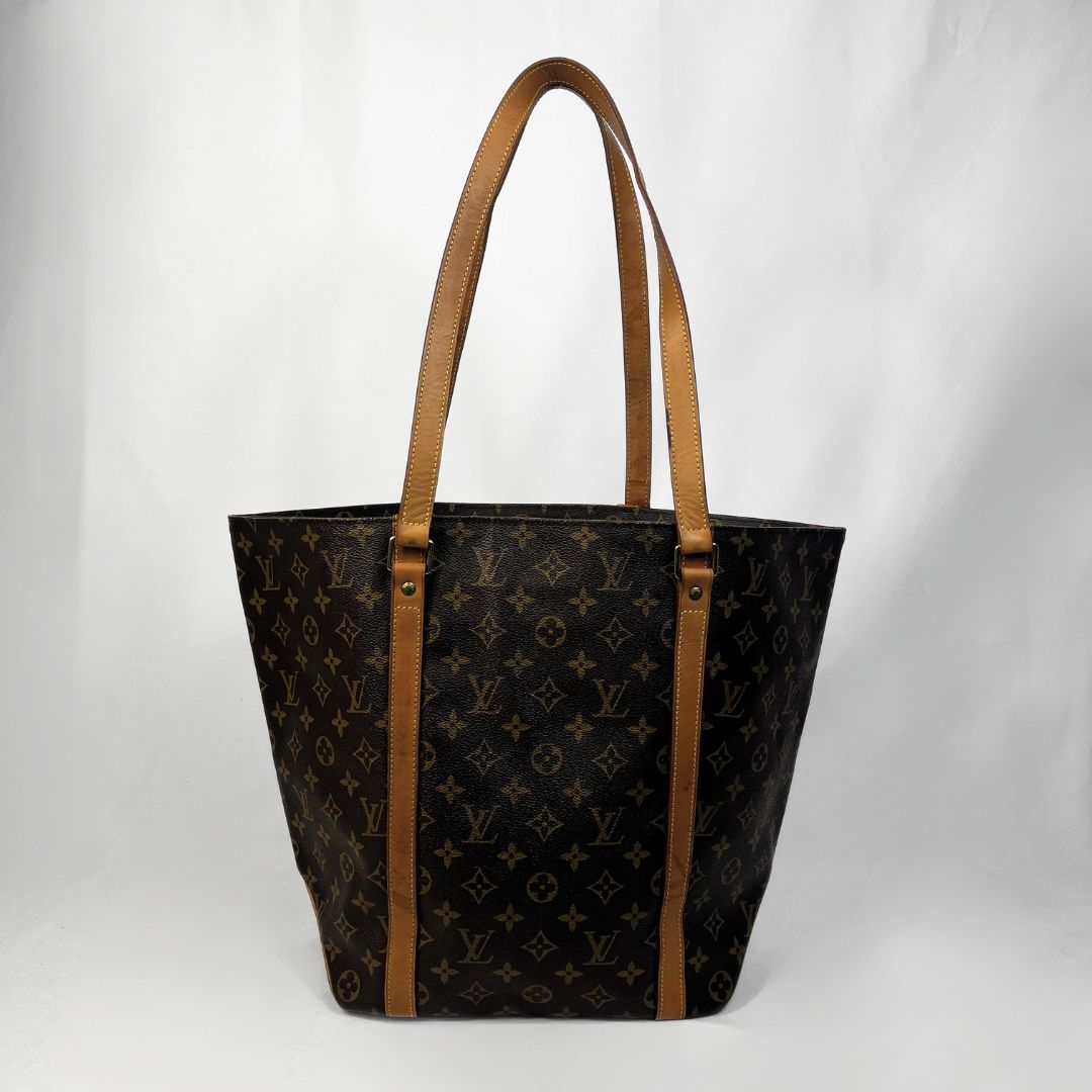 A Louis Vuitton monogram tote bag in dark brown coated canvas with tan leather shoulder straps and trim, photographed against a light gray fabric backdrop.