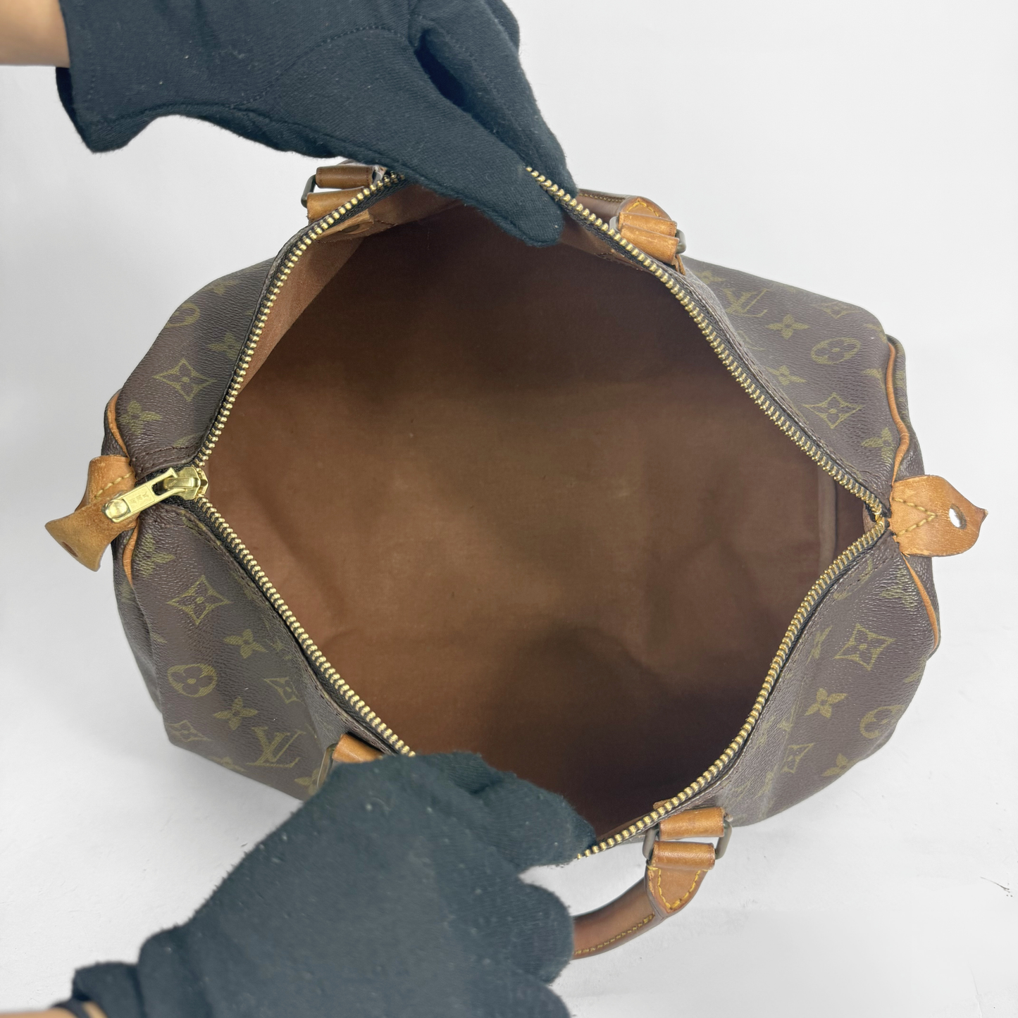 Ruskea Louis Vuitton kultaisella monogrammikuviolla varustettu laukku, jota pidetään auki hansikkaiden käsissä valkoisella taustalla