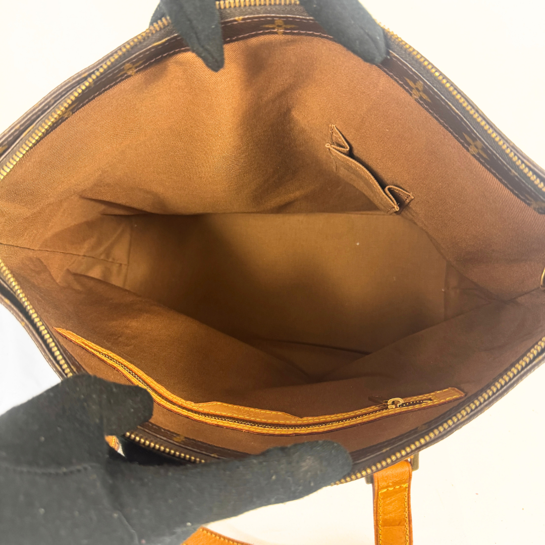 Open view of a Louis Vuitton handbag showing the brown monogram canvas exterior, tan interior lining, and gold-tone zipper. Gloved hands are holding the bag open against a white background.