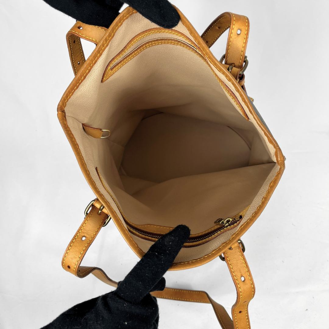 A tan leather handbag shown open from above, revealing a beige interior lining and a zippered compartment. Black-gloved hands are holding the bag open against a plain white background.