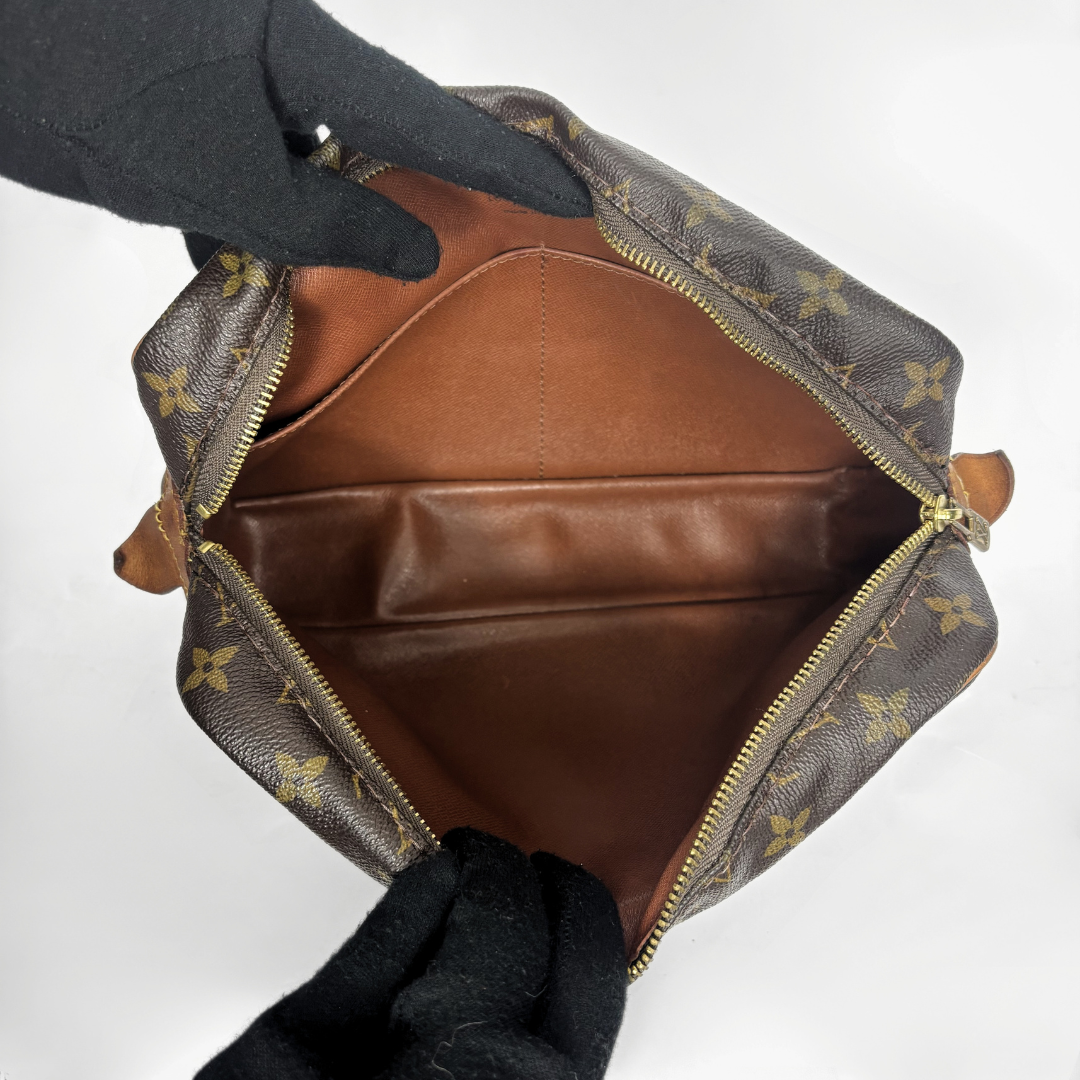 A Louis Vuitton monogram canvas bag shown open from above, revealing a brown leather interior, held by black-gloved hands against a plain white background.