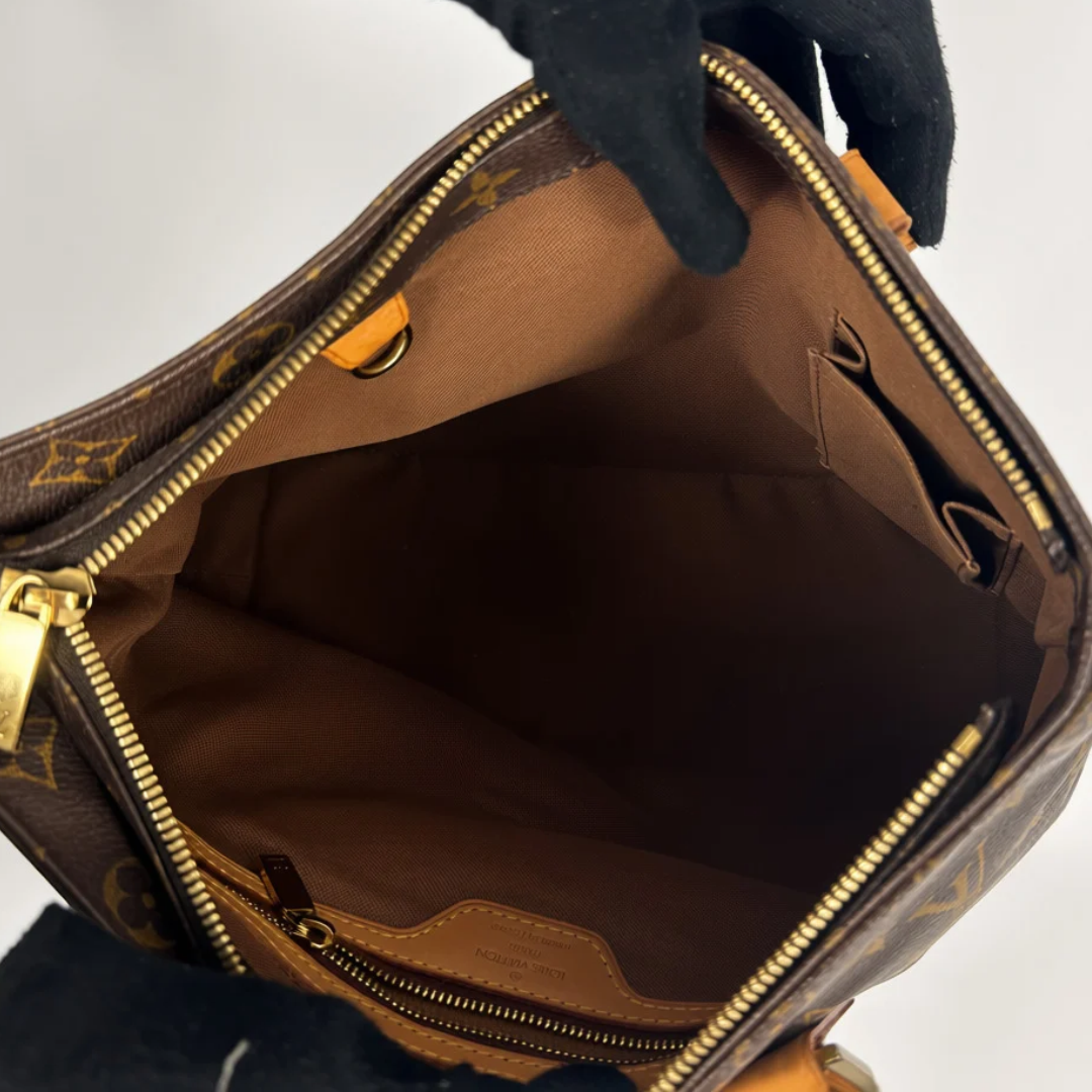 Louis Vuitton monogram canvas handbag shown open, with a black gloved hand holding the top zipper area. The interior is lined in brown and features gold-tone zippers and hardware.