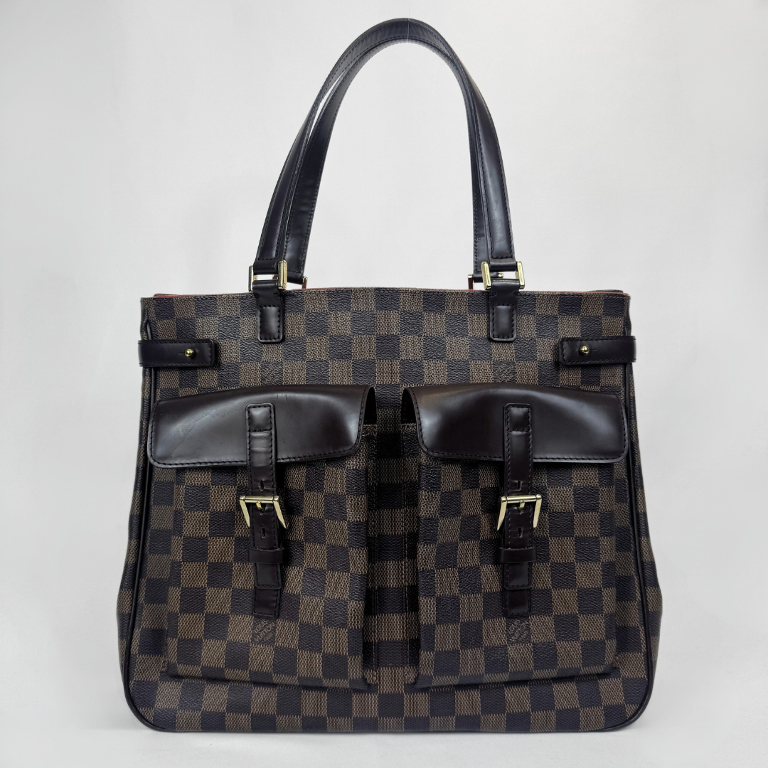 A Louis Vuitton tote bag in Damier Ebene canvas with dark brown leather handles and trim, featuring two front utility pockets with buckle details, set against a plain light gray background.