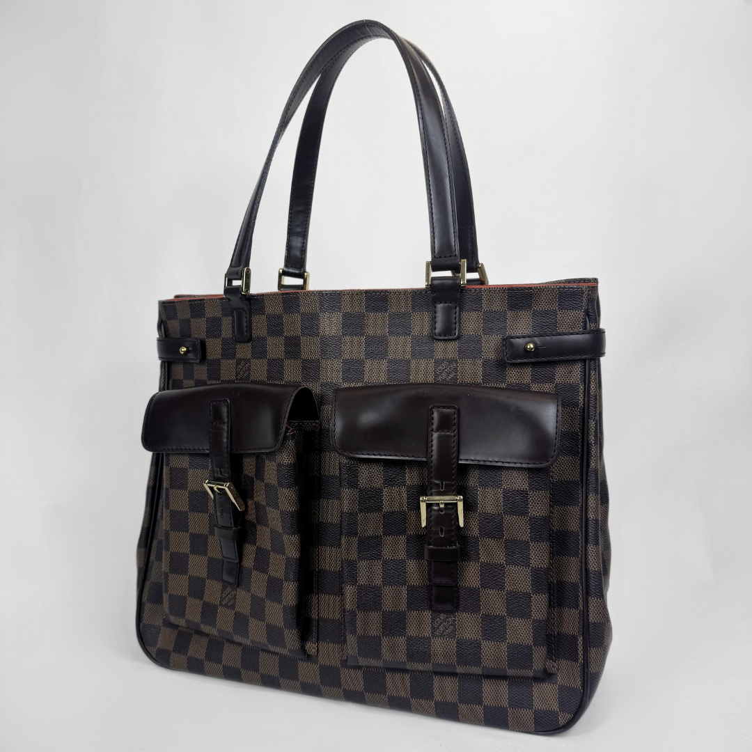A Louis Vuitton tote bag in the Damier Ebene checkered canvas with dark brown leather trim, dual top handles, and two front flap pockets with buckle details, photographed on a light gray background.