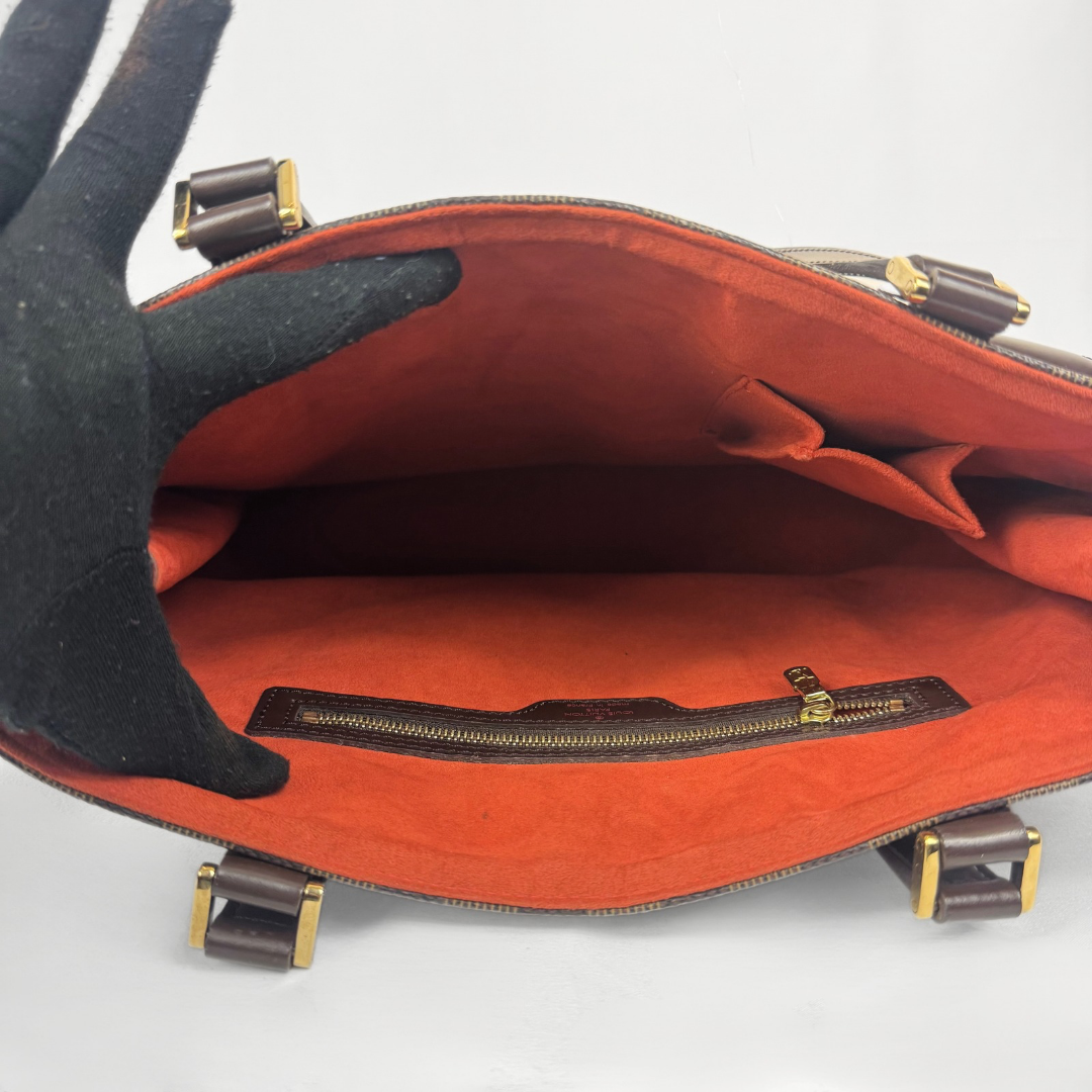 An open designer handbag with a bright orange-red suede interior, gold-tone hardware, and a black fabric glove holding the left side open against a white background.